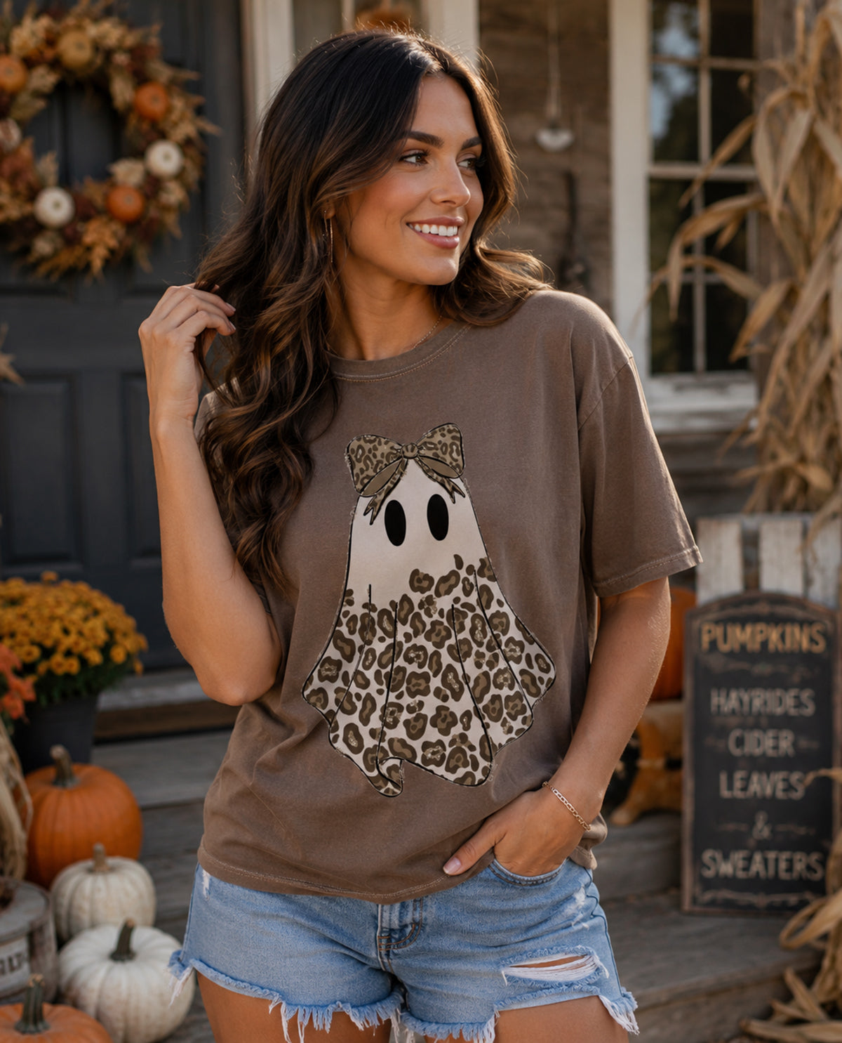 Woman wearing a brown t-shirt with a ghost design in front of a decorated house for Halloween.