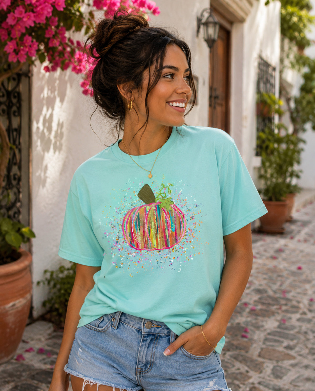 Woman wearing a light blue t-shirt with a colorful pumpkin design, standing on a street with plants and flowers.