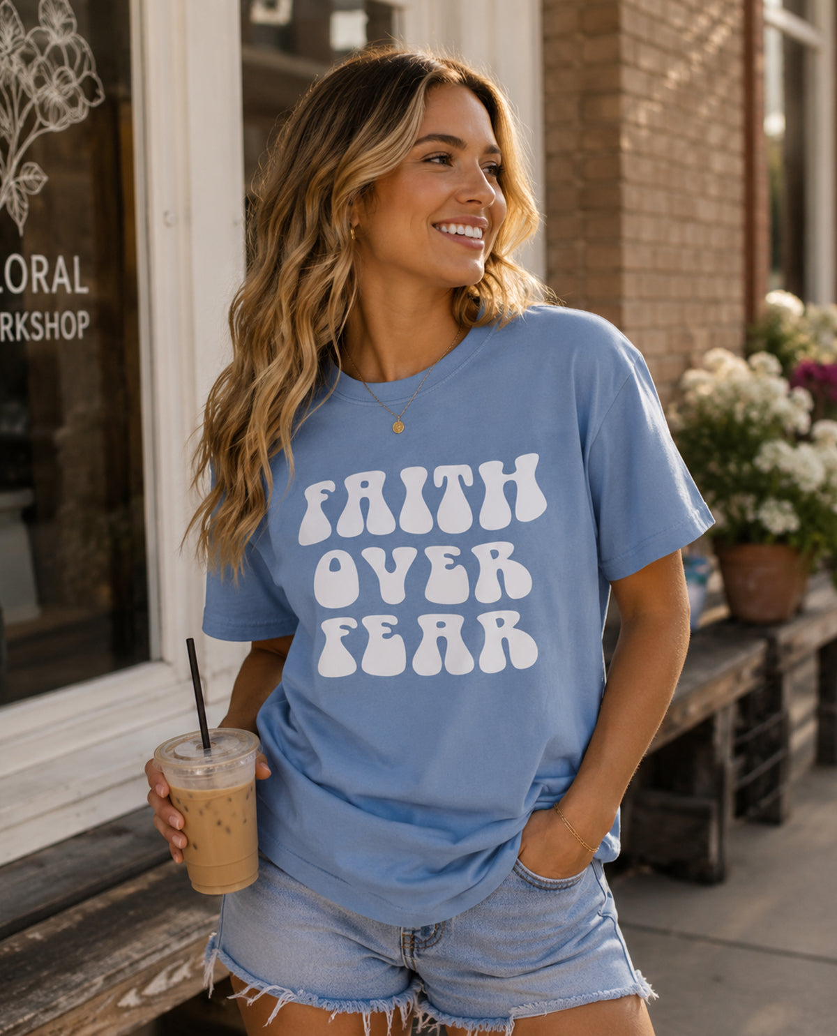 Woman wearing a blue t-shirt with 'Faith Over Fear' text, holding an iced coffee, standing outside a store.