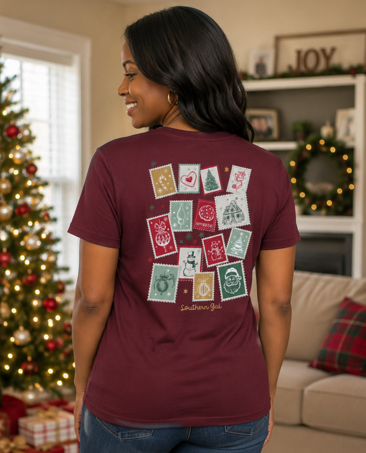 Woman wearing a maroon t-shirt with Christmas-themed designs in a living room decorated for the holidays.