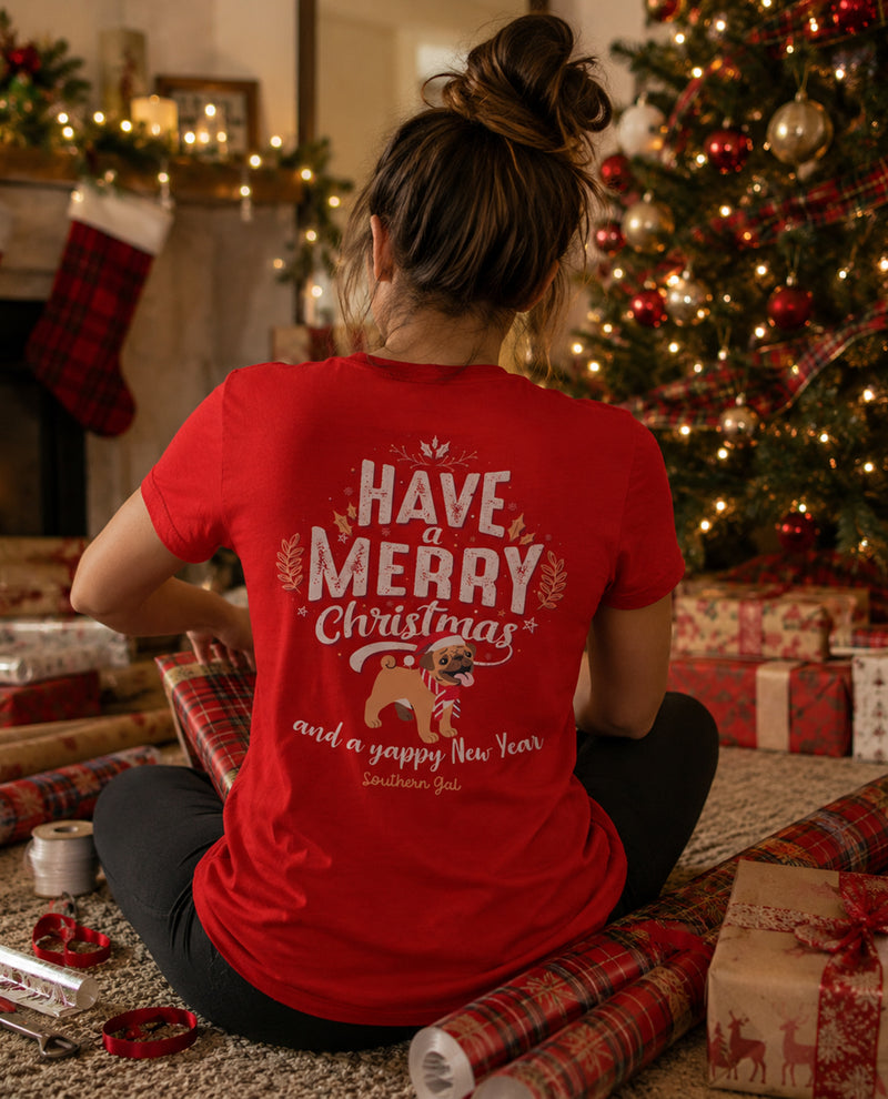 Person wearing a red 'Have a Merry Christmas and a Happy New Year' shirt in a festive setting with a Christmas tree and presents.