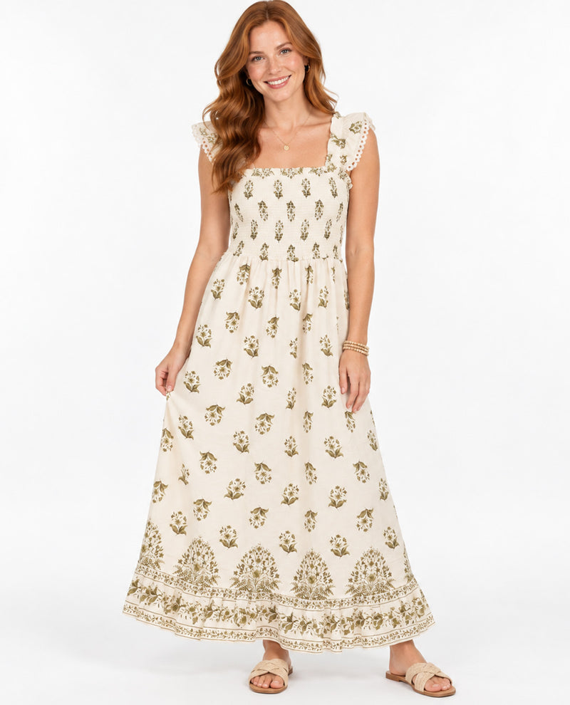 A woman modeling a sleeveless cream maxi dress with a small olive green floral block print and a decorative border along the hem, set against a plain white studio background.
