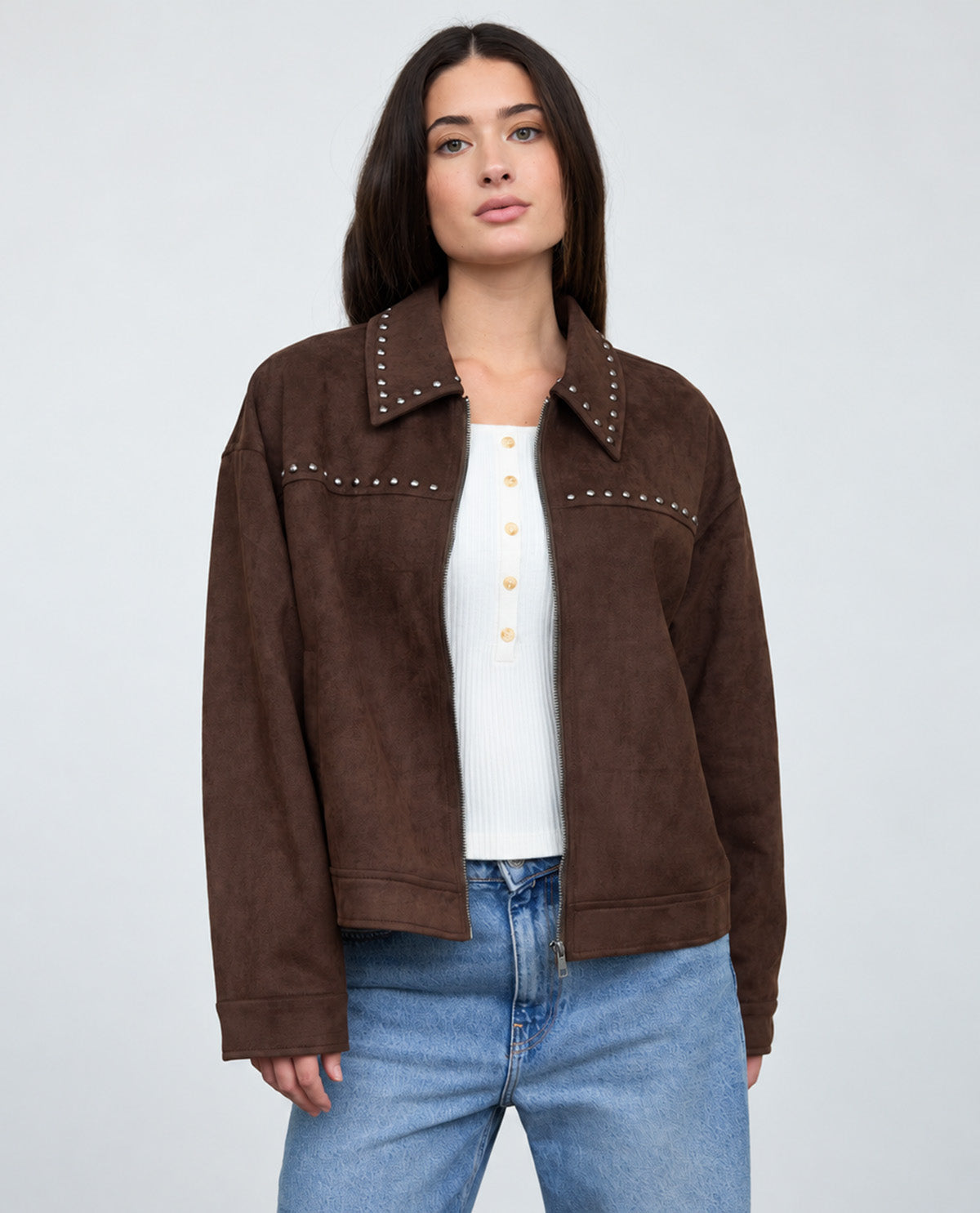 A woman modeling a brown suede jacket with silver stud detailing on the collar and chest, worn over a white henley top and blue jeans, against a light gray studio background.