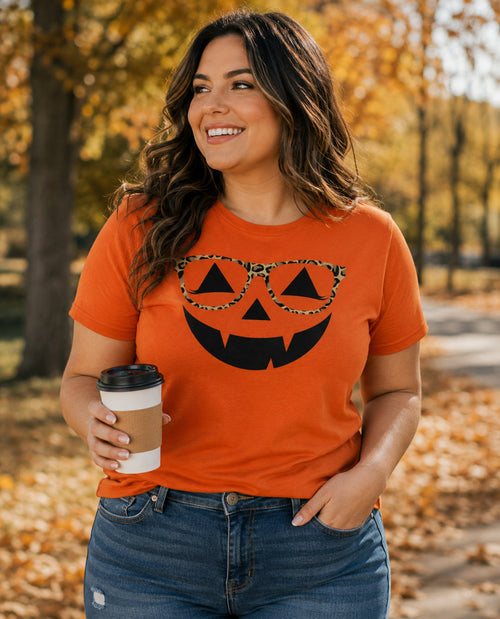 Woman wearing an orange t-shirt with a pumpkin design, standing outdoors with autumn foliage.