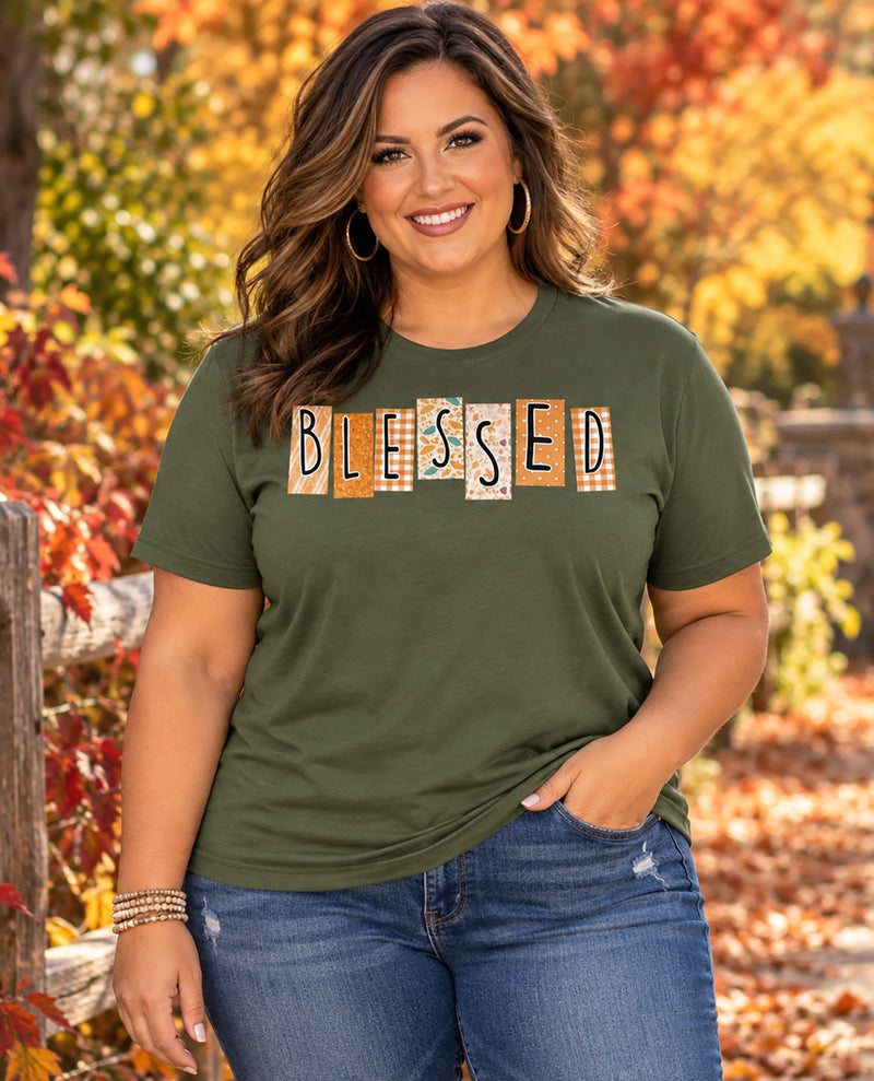 A woman wearing an olive green t-shirt with a 'BLESSED' patchwork graphic, paired with blue jeans, standing outdoors in front of a blurred autumn background of orange and yellow leaves.