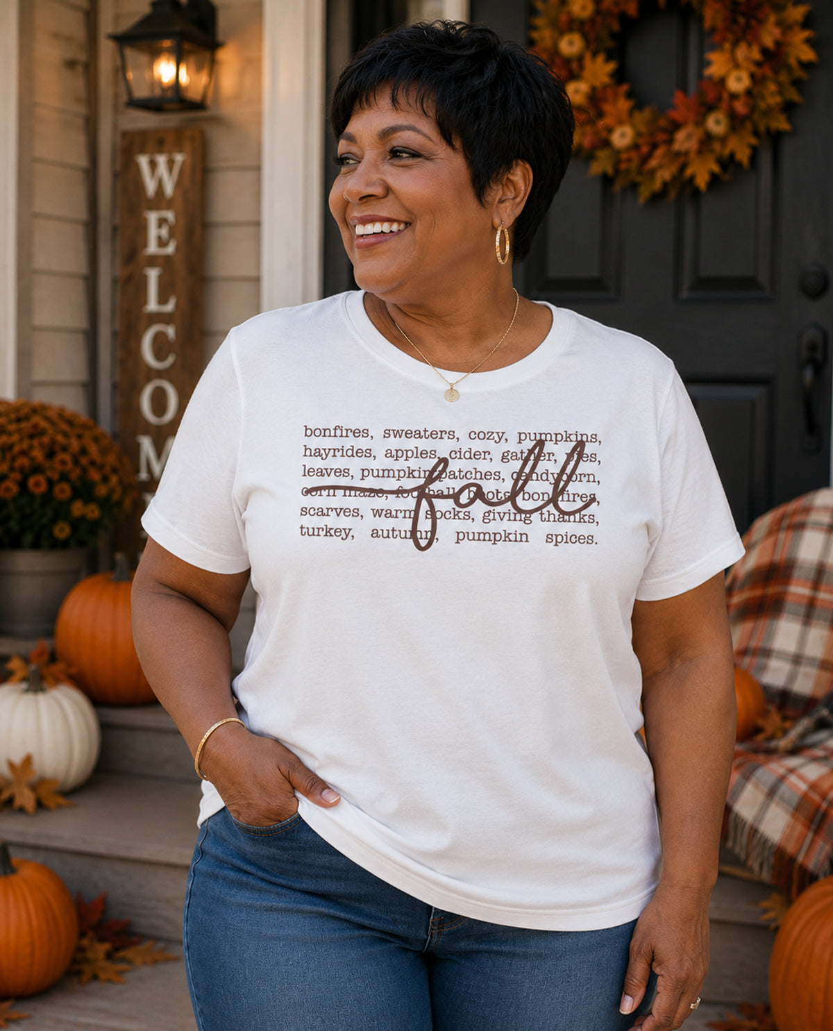 Woman wearing a white t-shirt with 'fall' definition print, standing in front of a decorated door with pumpkins and a wreath.