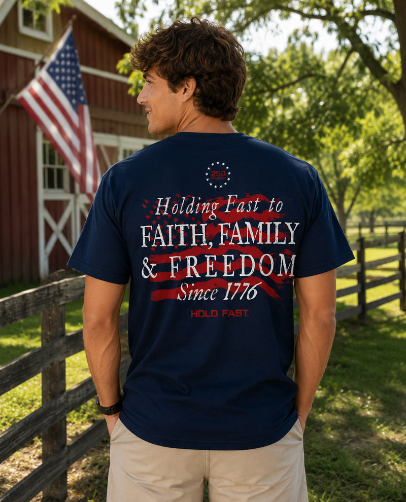 Man wearing a navy blue t-shirt with patriotic text in a rural setting with a red barn and American flag.