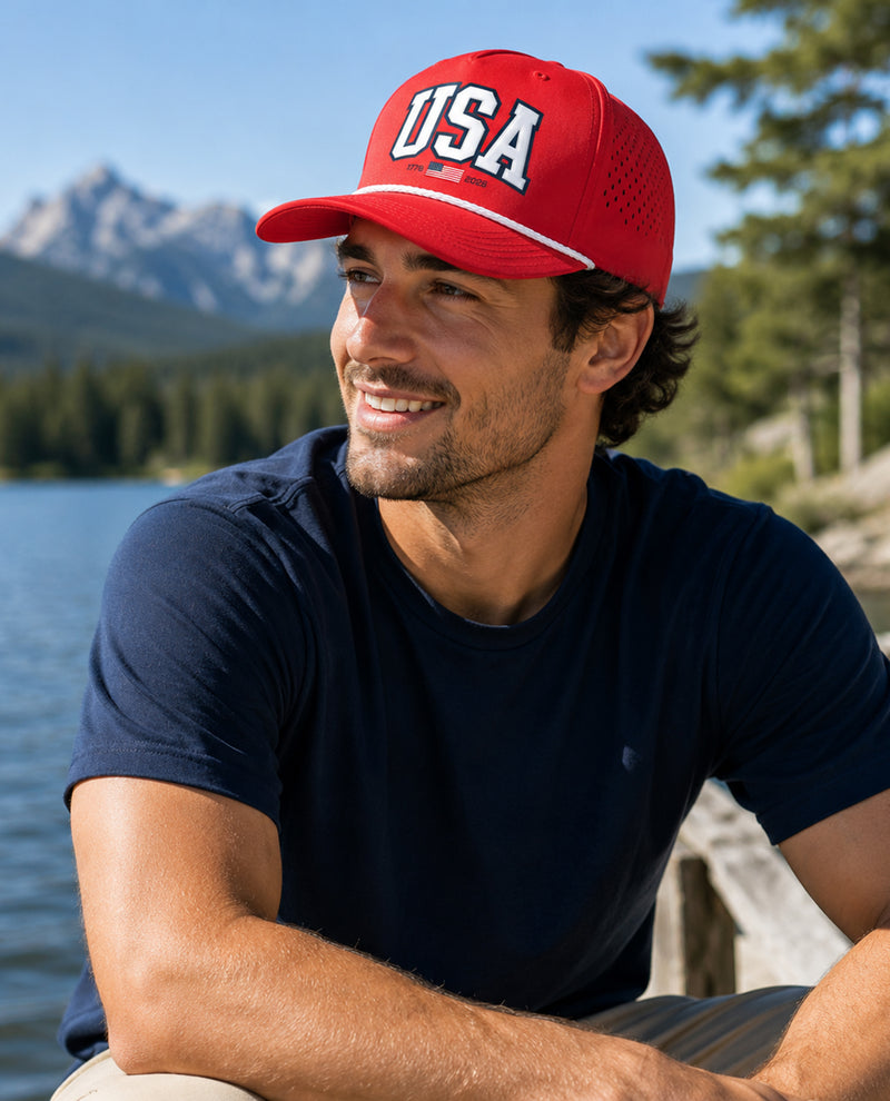 Man wearing a red 'USA' cap by a lake with mountains in the background