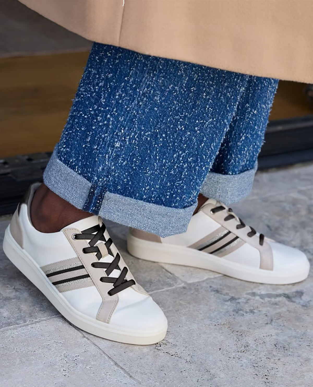 White low-top sneakers with black laces and beige side stripes, worn with cuffed blue jeans and a beige coat, shown on a person standing on a stone floor.