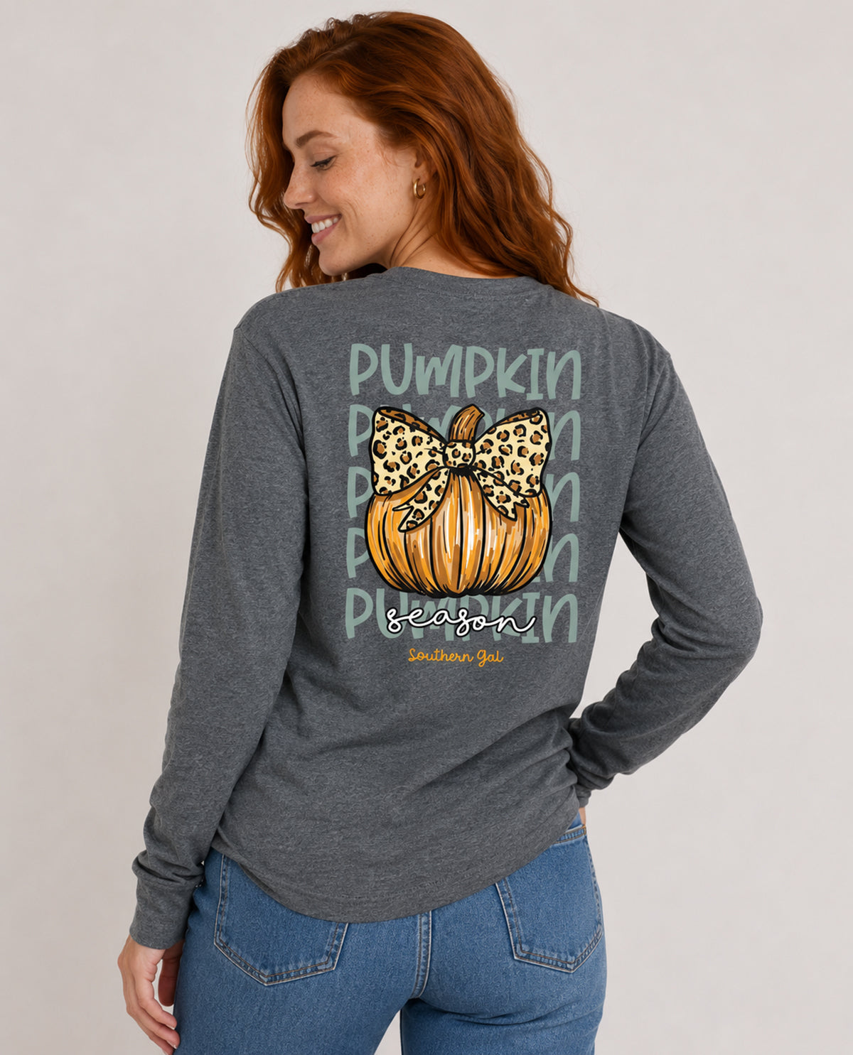 A woman wearing a dark heather gray long-sleeve shirt with a graphic on the back featuring a pumpkin, leopard print bow, and the words 'Pumpkin Season' and 'Southern Girl', paired with blue jeans against a light gray studio background.