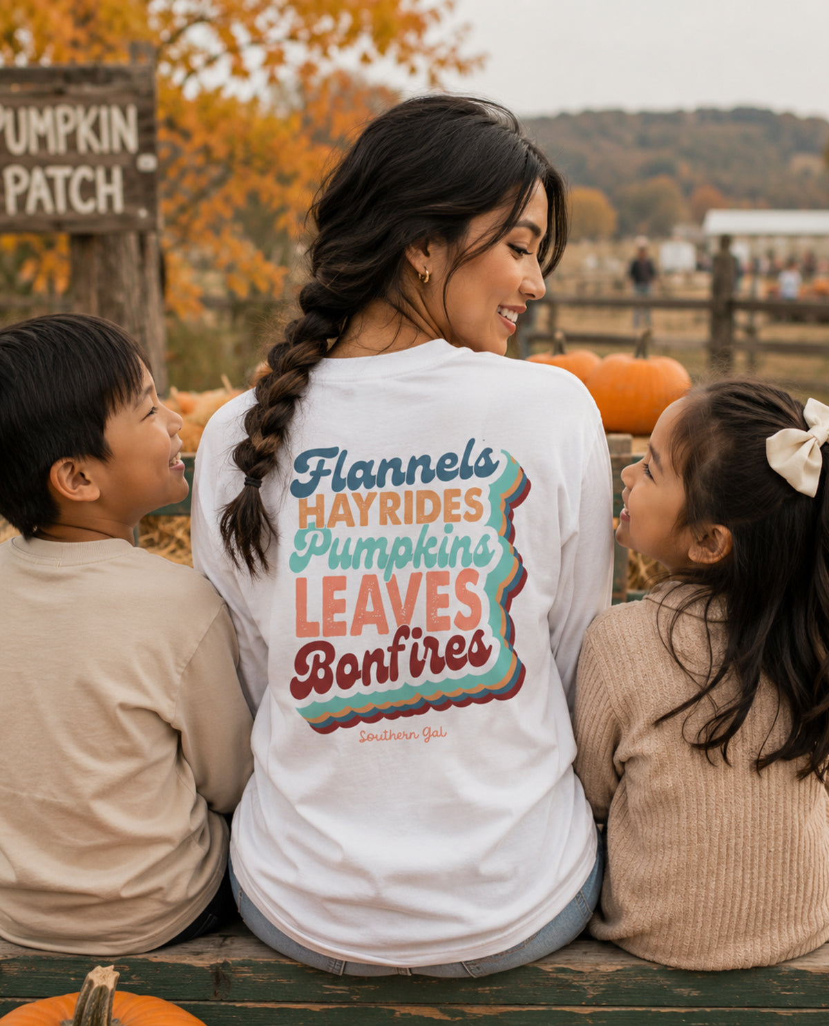 A woman wearing a white long-sleeve graphic t-shirt sits on a wooden bench outdoors with two children. The shirt features a colorful autumn-themed graphic reading 'Flannels Hayrides Pumpkins Leaves Bonfires' on the back.