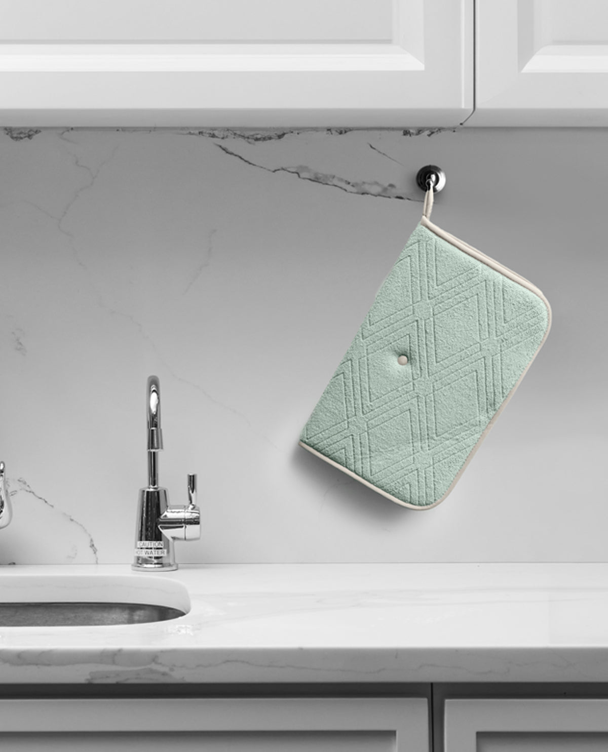 A mint green quilted potholder with a cream border hangs from a black hook on a white marble kitchen backsplash above a sink.