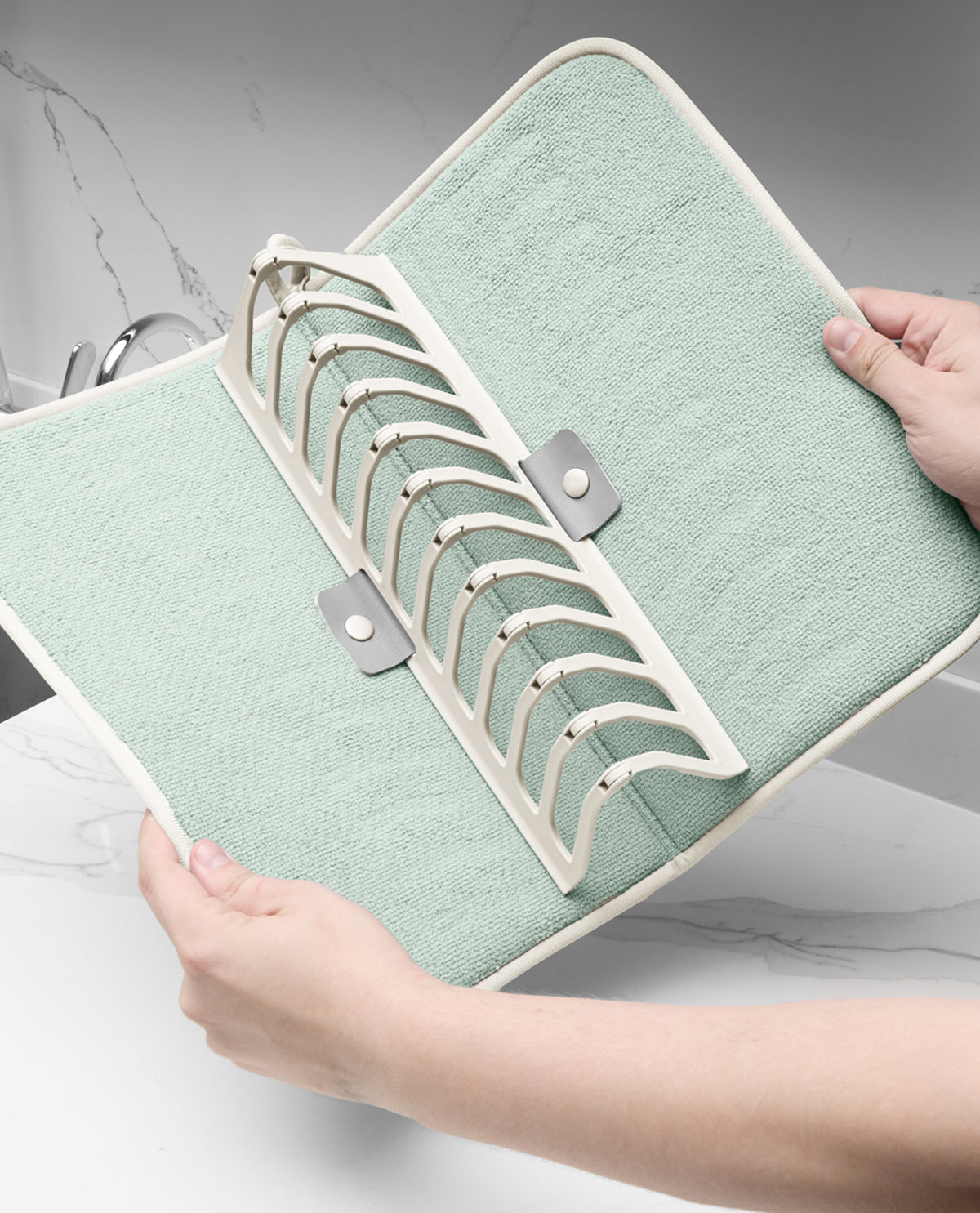 A mint green and off-white kitchen towel rack organizer with a curved design, held open by two hands over a white marble countertop.
