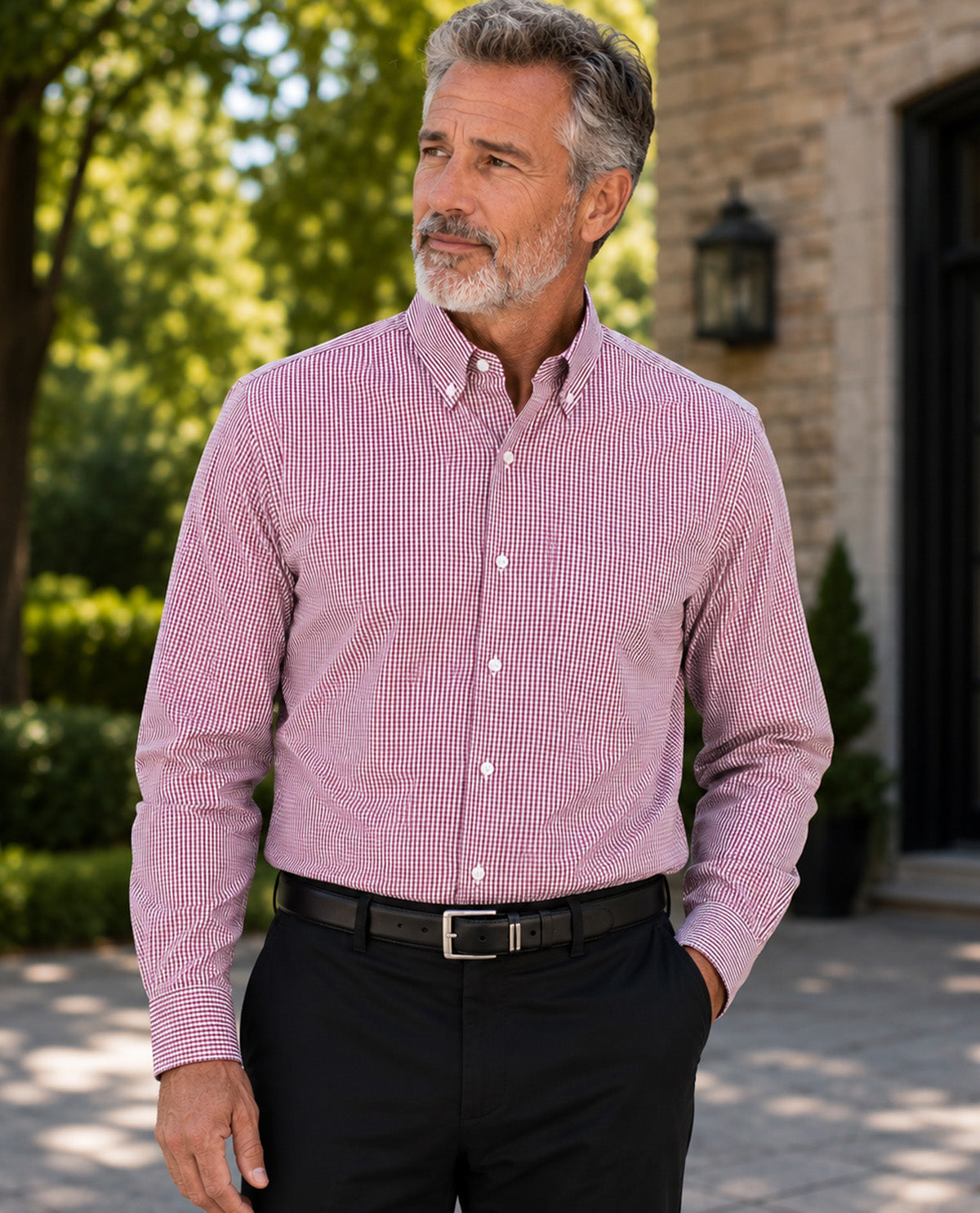 A man wearing a pink and white micro-checkered long-sleeve button-down shirt, paired with black trousers and a black belt, standing outdoors in front of a stone building.