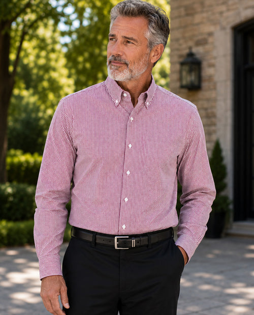 A man wearing a pink and white micro-checkered long-sleeve button-down shirt, paired with black trousers and a black belt, standing outdoors in front of a stone building.