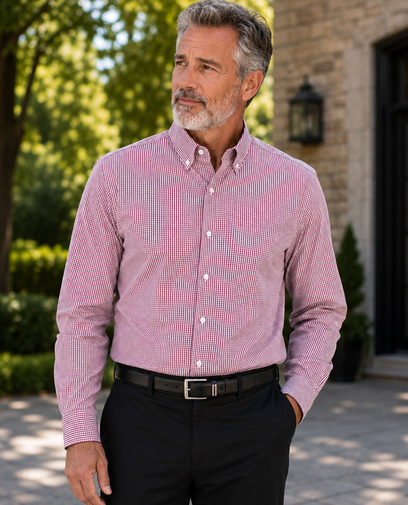 A man wearing a pink and white micro-checkered long-sleeve button-down shirt, paired with black trousers and a black belt, standing outdoors in front of a stone building.