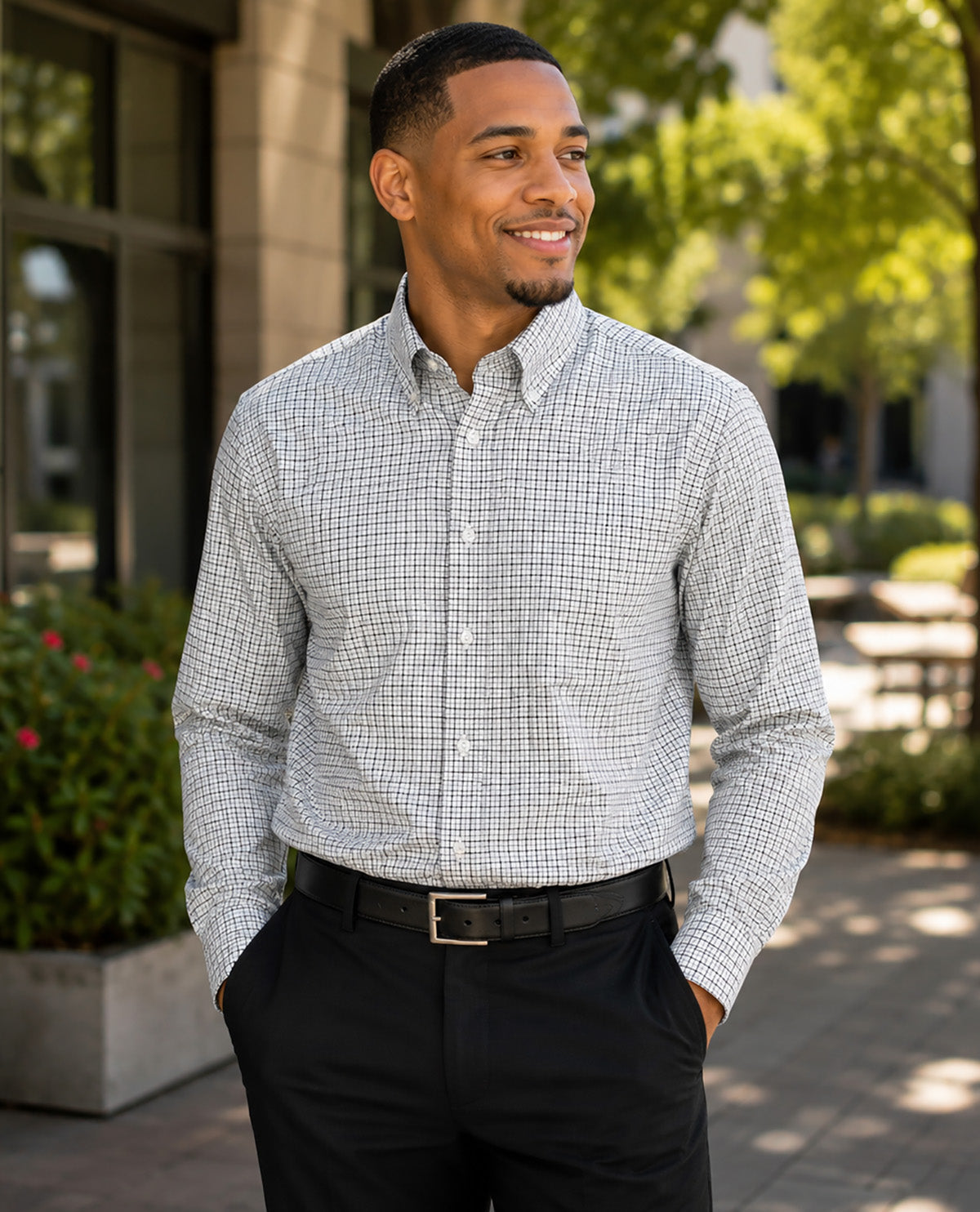 A man wearing a light gray and white micro-check patterned button-down shirt, paired with black dress pants and a black belt, standing outdoors in front of a modern building with green trees in the background.