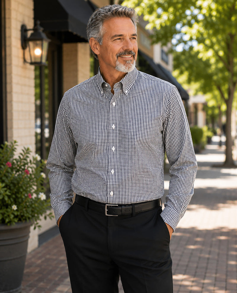 A man wearing a long-sleeve button-down shirt with a small black and white gingham pattern, paired with black trousers and a black belt, standing on a sunny brick sidewalk.