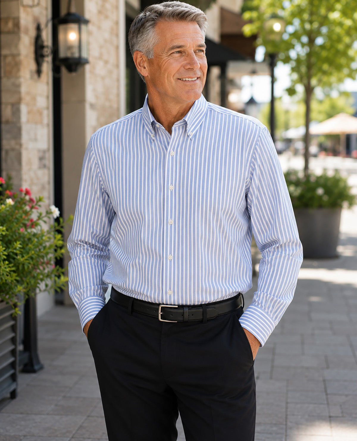 A smiling older man standing outdoors on a paved walkway, wearing a light blue and white vertically striped button-down dress shirt with black trousers and a black belt.