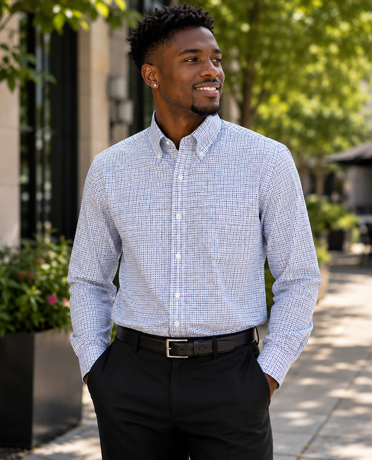 A man wearing a light blue and white micro-check patterned button-down shirt, paired with black dress pants and a black belt, standing outdoors on a paved walkway with green trees in the background.