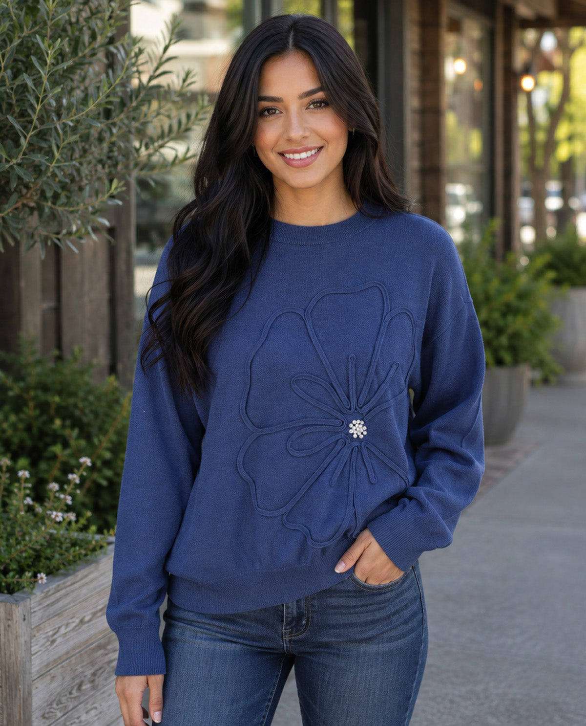 A woman wearing a navy blue crewneck sweatshirt with a large floral applique and a rhinestone center, paired with blue jeans, standing outdoors on a sidewalk.