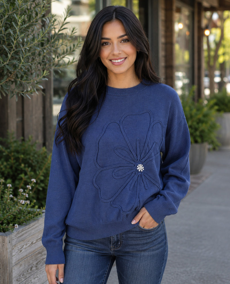 A woman wearing a navy blue crewneck sweatshirt with a large floral applique and a rhinestone center, paired with blue jeans, standing outdoors on a sidewalk.