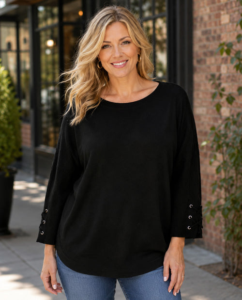 A smiling blonde woman wearing a black long-sleeve top with decorative silver grommets on the cuffs, paired with blue jeans, standing outdoors in front of a brick building.