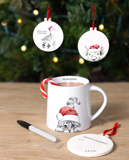 White mug with cat design, candy cane, and Christmas ornaments on a wooden surface with blurred Christmas tree background.
