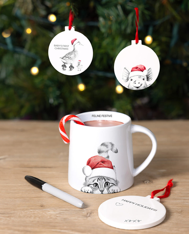 White mug with cat design, candy cane, and Christmas ornaments on a wooden surface with blurred Christmas tree background.