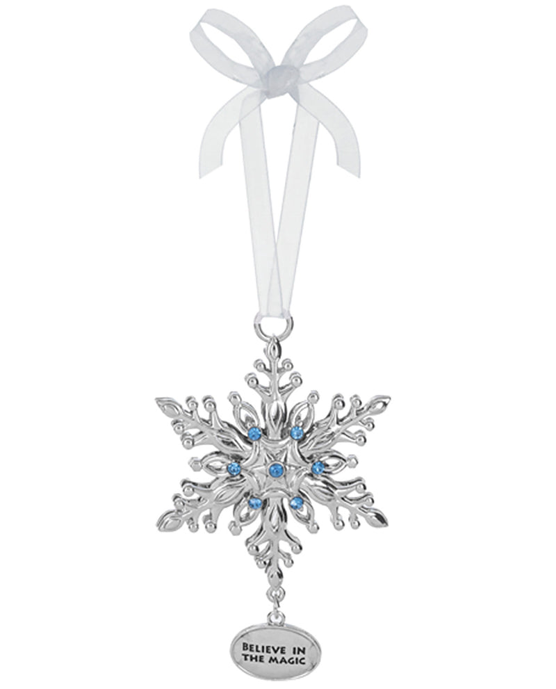 A silver snowflake Christmas ornament with small blue rhinestones in the center, hanging from a white ribbon bow. A small oval tag at the bottom reads 'BELIEVE IN THE MAGIC'.