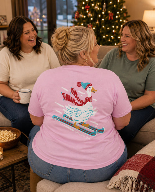Woman wearing a pink t-shirt with a goose graphic, sitting on a couch with two other people in a cozy living room.