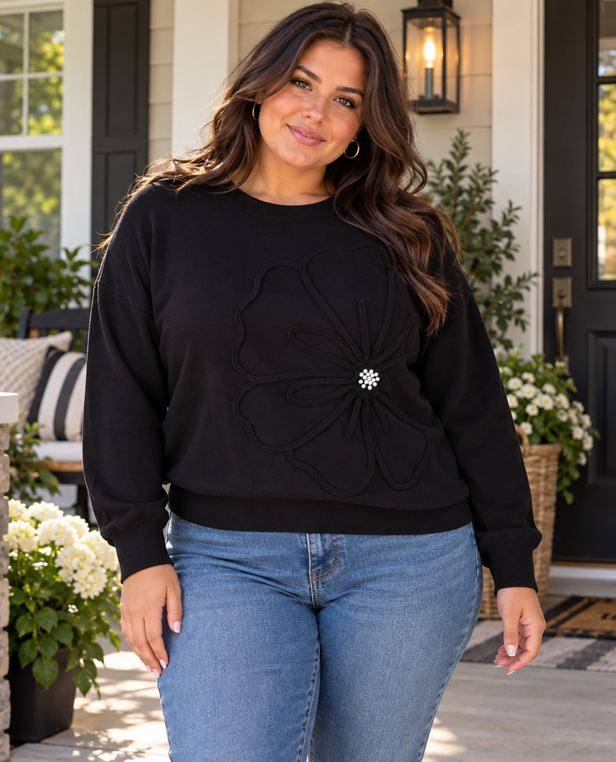 A woman wearing a black crewneck sweatshirt with a large 3D floral applique and a small silver rhinestone center, paired with blue jeans, standing on a front porch.