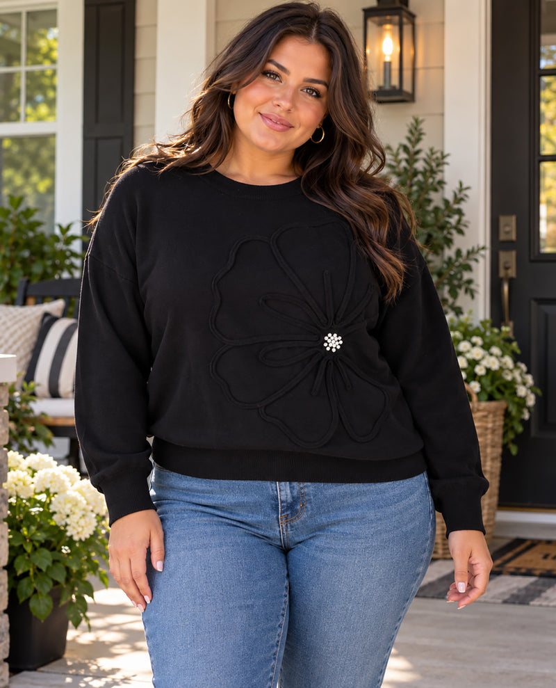 A woman wearing a black crewneck sweatshirt with a large 3D floral applique and a small silver rhinestone center, paired with blue jeans, standing on a front porch.