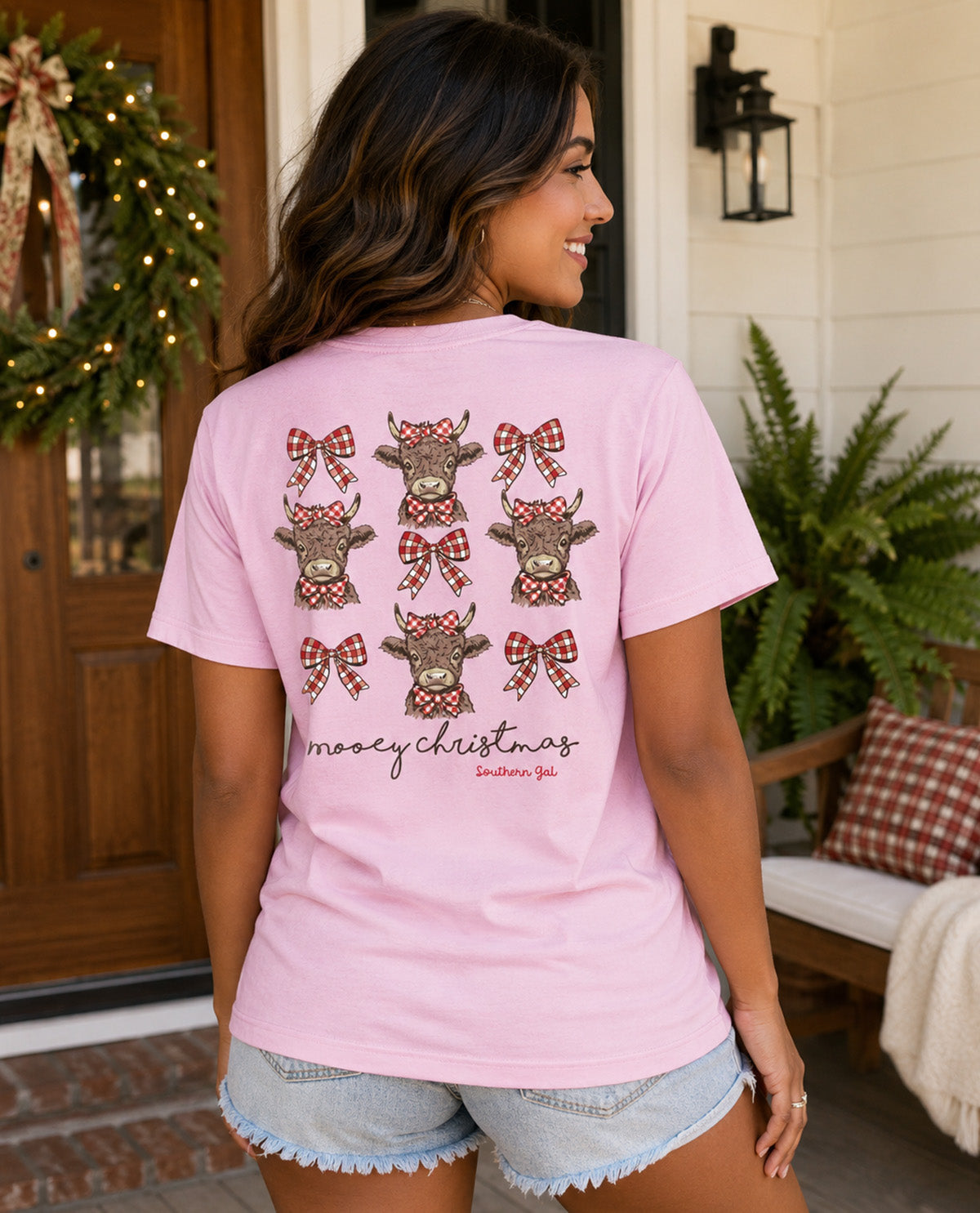 A woman shown from the back wearing a light pink t-shirt with cows wearing Christmas bows, paired with denim shorts, standing in front of a decorated front porch.
