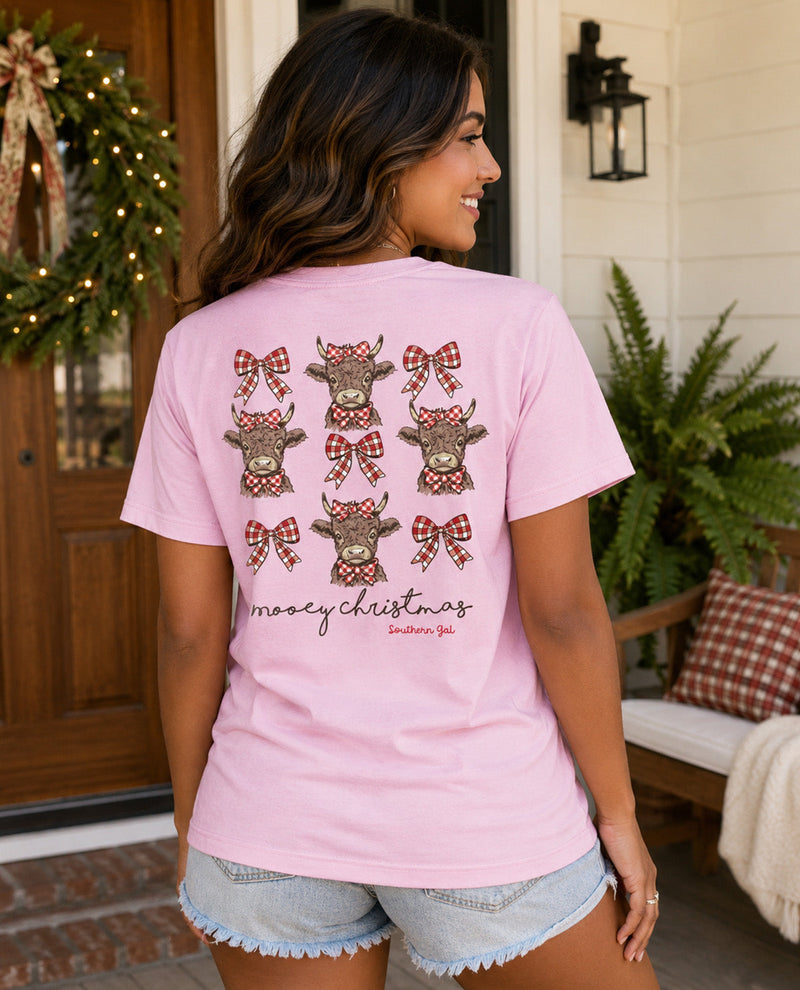 A woman shown from the back wearing a light pink t-shirt with cows wearing Christmas bows, paired with denim shorts, standing in front of a decorated front porch.