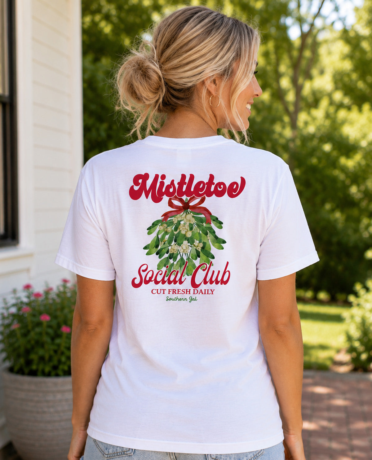 A woman viewed from behind wearing a white graphic t-shirt with a red and green mistletoe design and the text 'Mistletoe Social Club' on the back, set against an outdoor garden background.