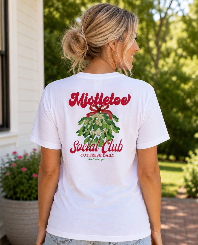 A woman viewed from behind wearing a white graphic t-shirt with a red and green mistletoe design and the text 'Mistletoe Social Club' on the back, set against an outdoor garden background.