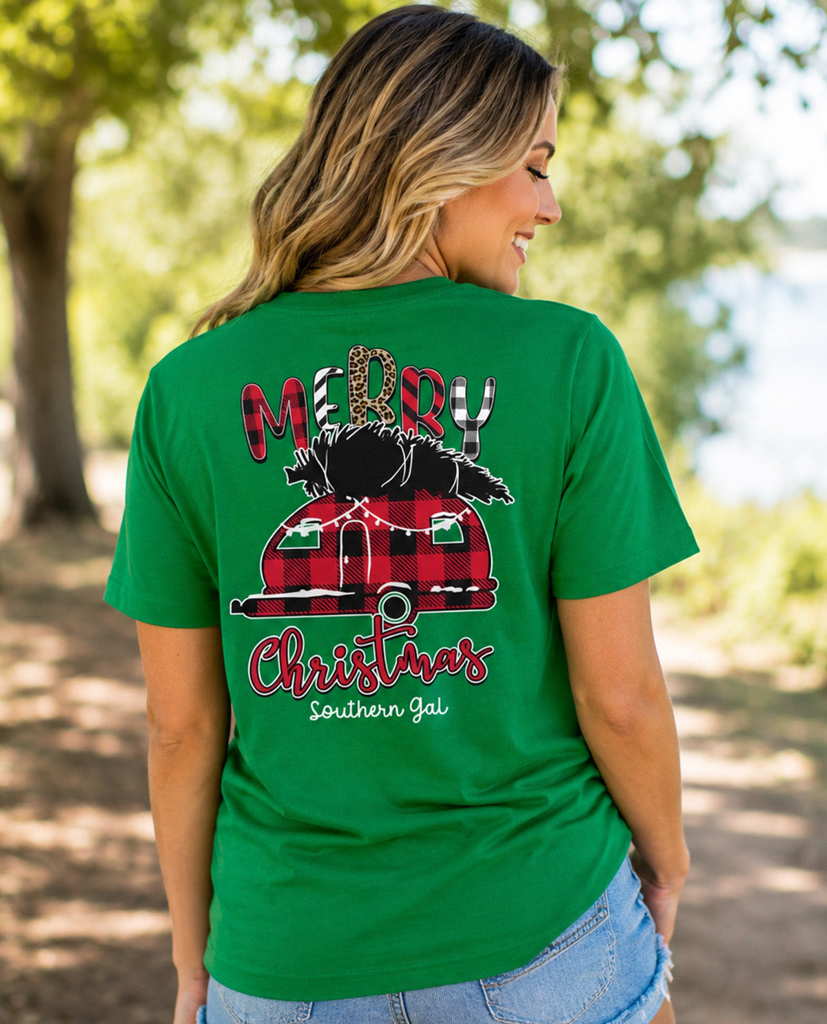 A woman wearing a green short-sleeve t-shirt with a large Christmas graphic on the back, photographed outdoors in a park setting.
