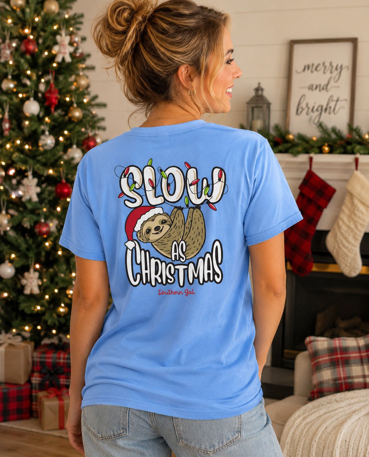A woman shown from behind wearing a light blue graphic t-shirt with a Christmas sloth design and the text 'SLOW AS CHRISTMAS' in a cozy holiday living room setting.