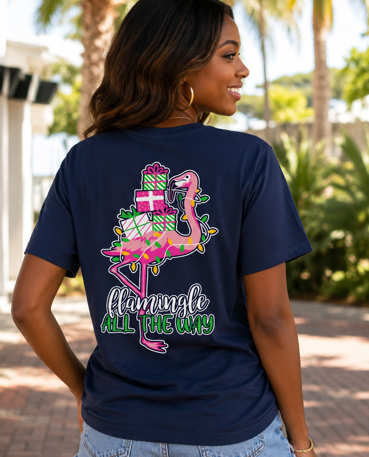A woman shown from the back wearing a navy blue t-shirt with a colorful flamingo graphic carrying wrapped presents and the text 'FLAMINGLE ALL THE WAY'.