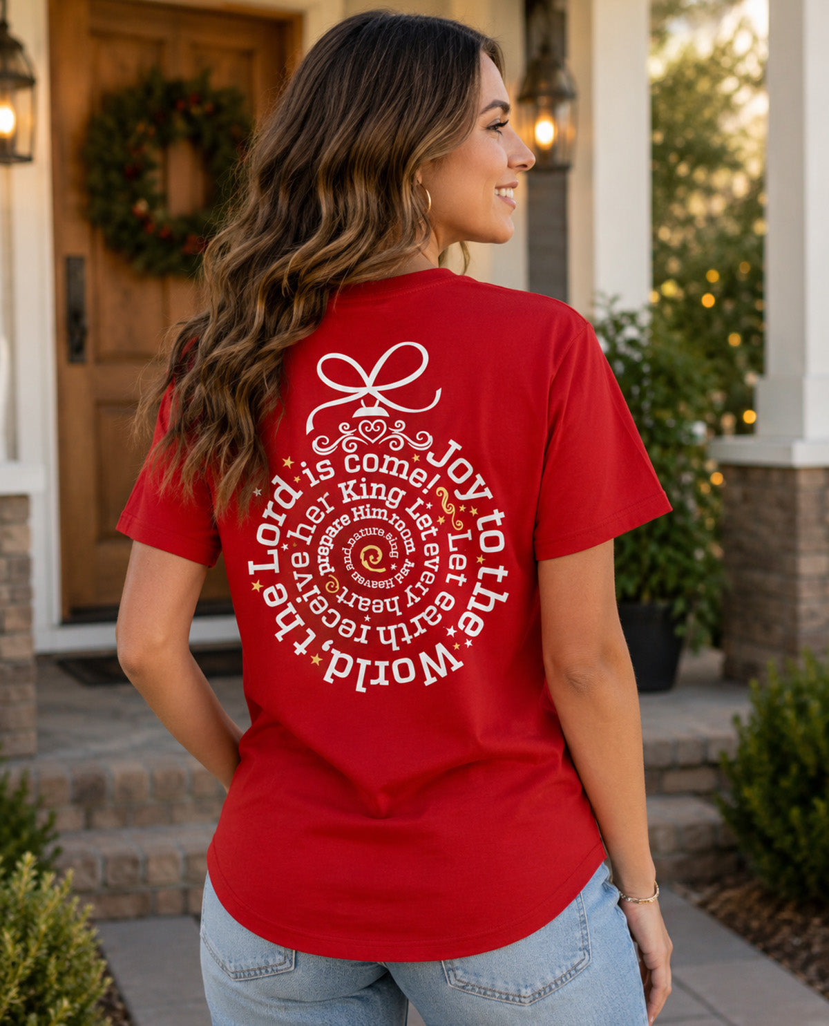A woman wearing a red short-sleeve t-shirt with a large white circular Christmas-themed graphic on the back, standing in front of a house porch.