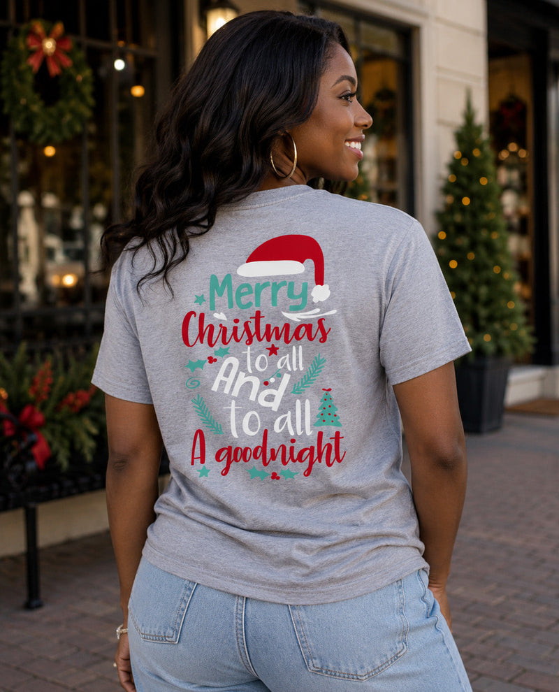 A woman wearing a heather gray t-shirt with a festive Christmas graphic on the back, standing outdoors in front of a storefront with Christmas decorations.