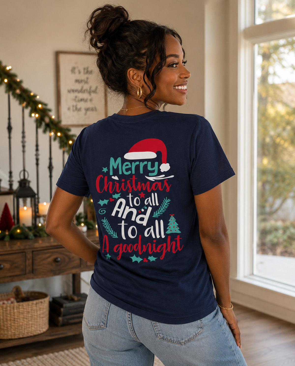 A woman wearing a navy blue graphic t-shirt with a festive Christmas design on the back, posing in a warmly lit indoor room.