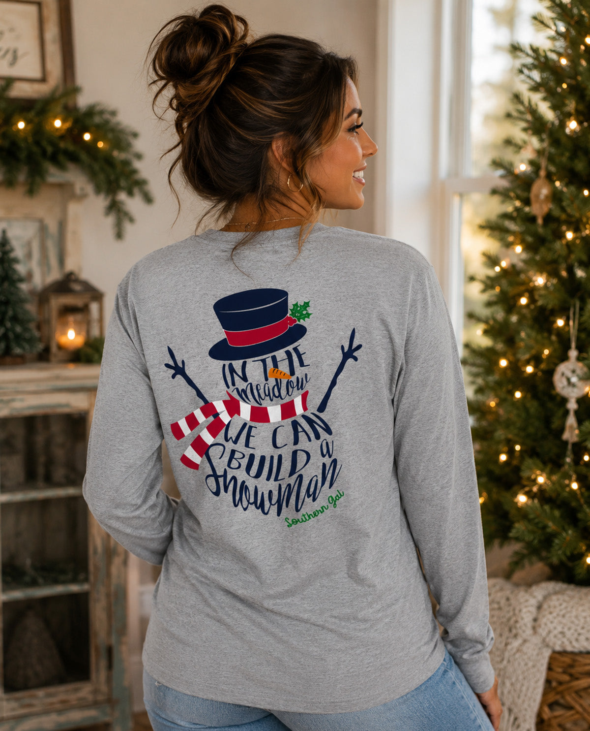 A woman wearing a heather gray long-sleeve t-shirt, viewed from behind. The shirt features a large Christmas graphic on the back with a top hat, snowman, scarf, and the text 'IN THE MEADOW WE CAN BUILD A SNOWMAN' with 'Southern Gal' written below.
