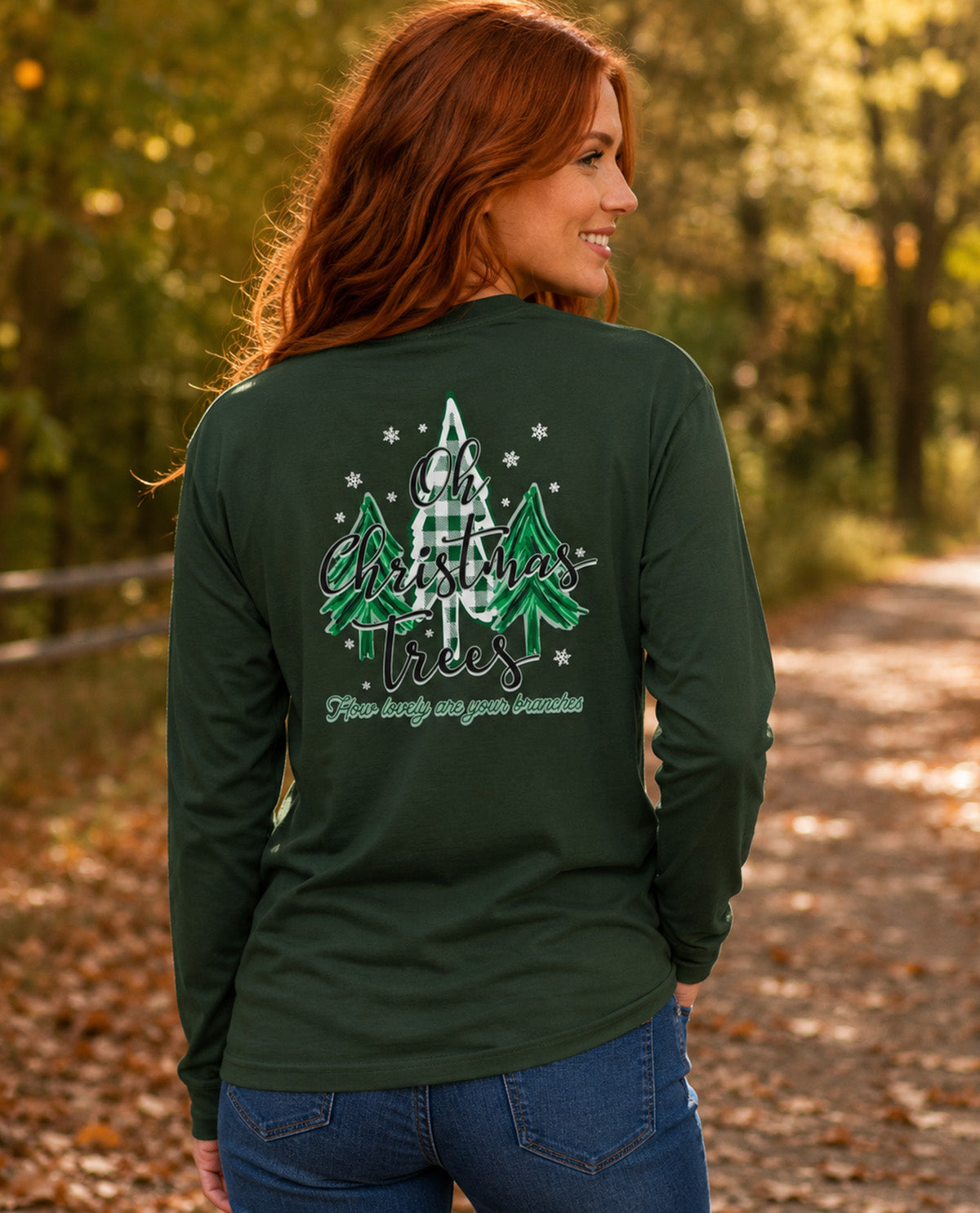 A woman with red hair is shown from behind wearing a dark forest green long-sleeve shirt with a graphic on the back. The graphic features green and white plaid Christmas trees, white snowflakes, and the text 'Oh Christmas Trees' .