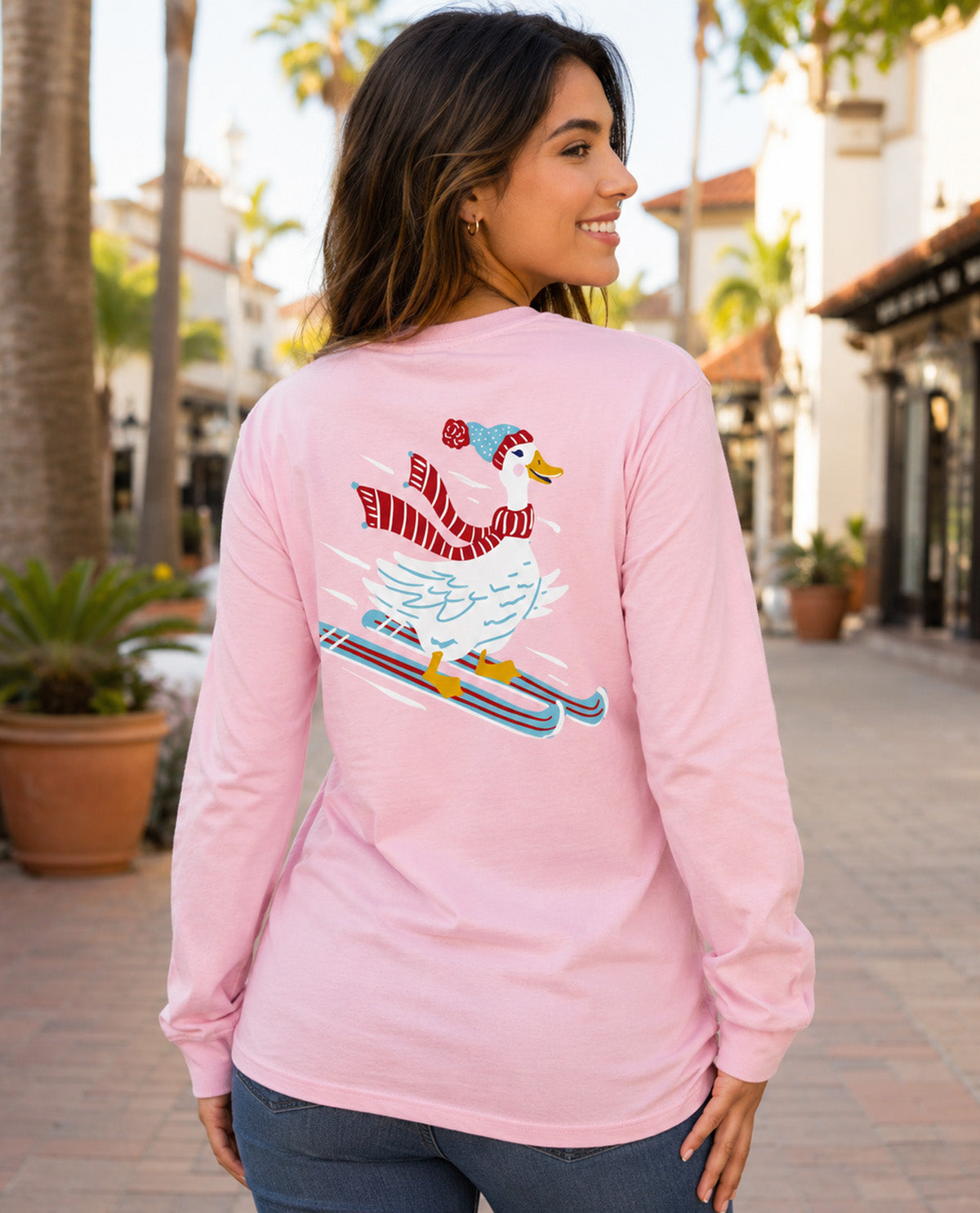 A woman wearing a light pink long-sleeve t-shirt with a graphic of a goose skiing on the back, set against a blurred outdoor storefront background.