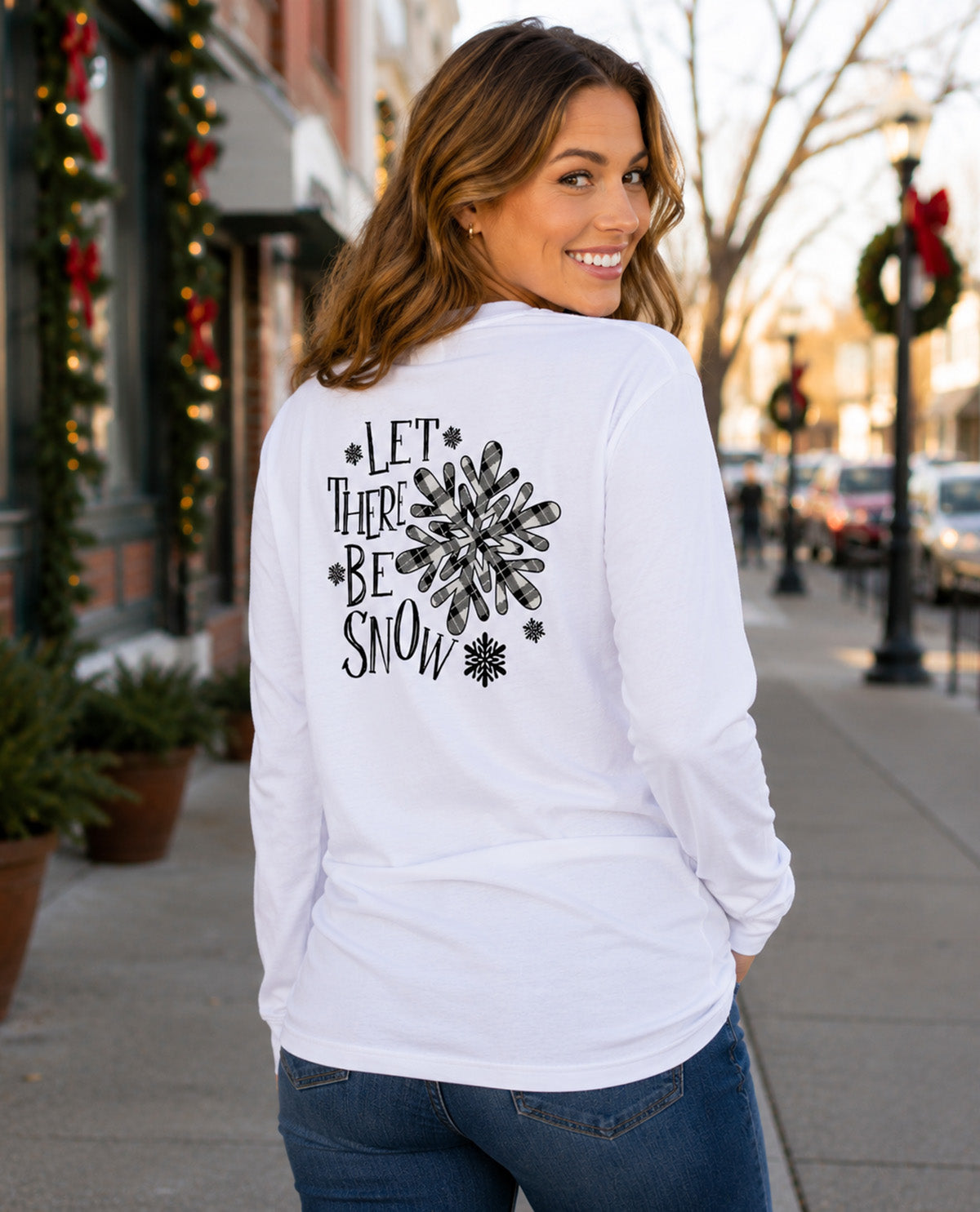 A woman wearing a white long-sleeve t-shirt with a black graphic print that reads 'LET THERE BE SNOW' with a snowflake design, standing on a city sidewalk.