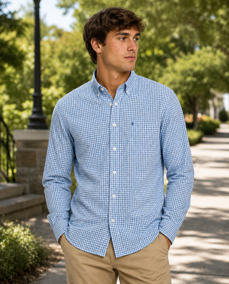 A man wearing a light blue and white gingham long-sleeve button-down shirt with khaki pants, standing outdoors on a sunny path with trees in the background.