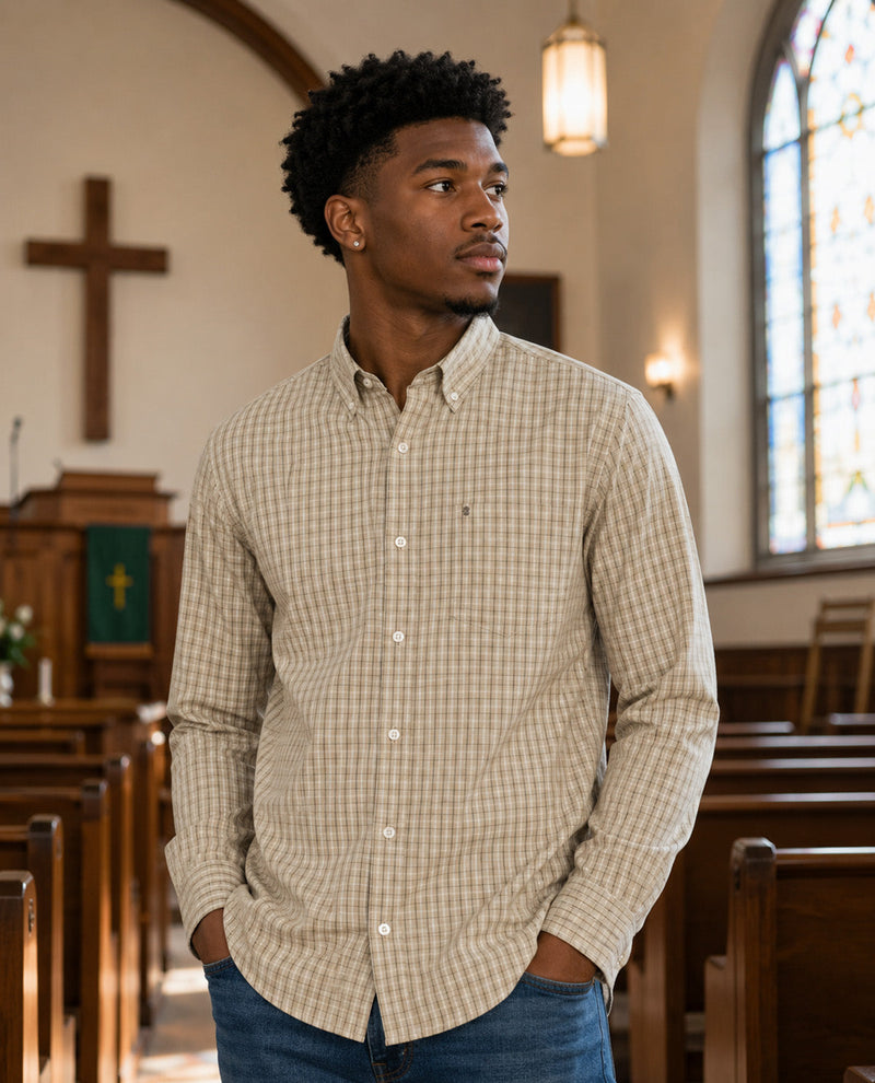 A man wearing a beige and brown checkered button-down shirt with blue jeans, standing in a church aisle with wooden pews and a stained glass window in the background.