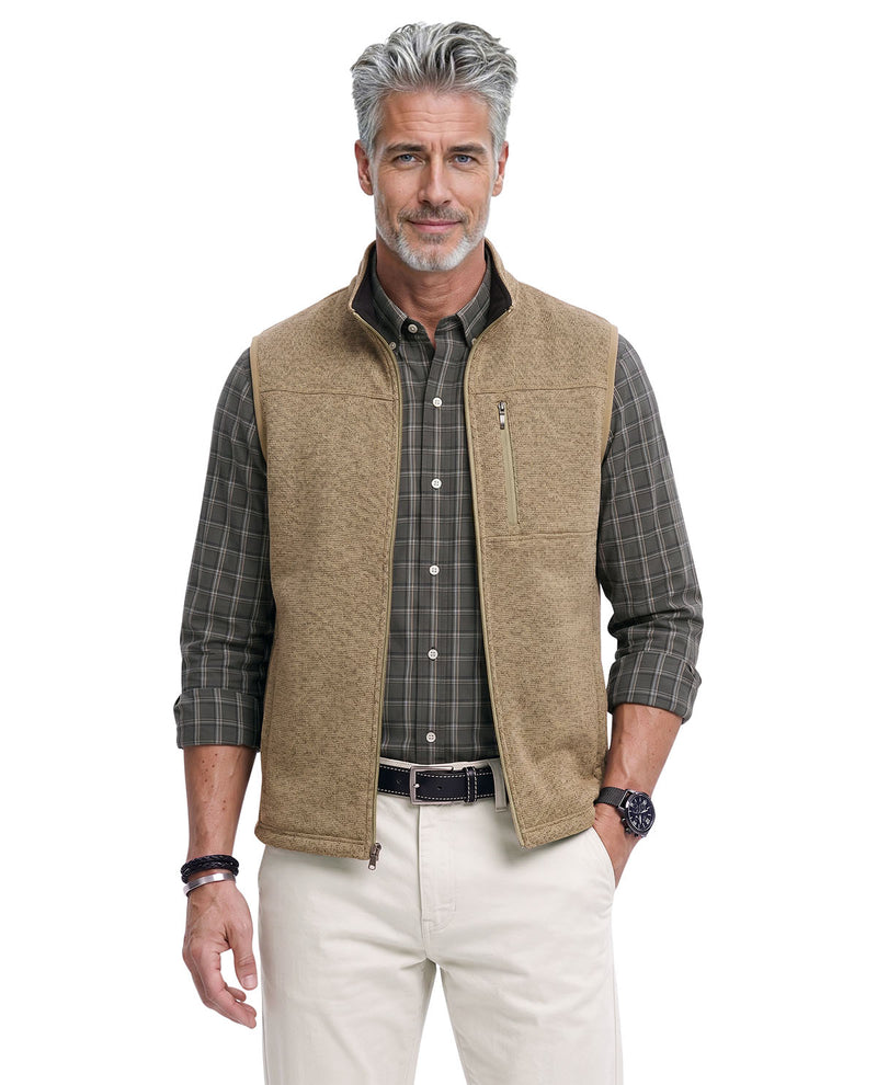 A middle-aged man with gray hair and a beard wearing a tan fleece vest over a dark green plaid button-down shirt, paired with cream pants, against a plain white background.